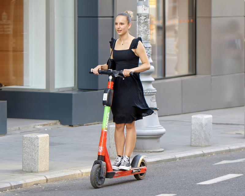 30 - Young woman wearing a stylish black dress paired with classic high-top sneakers while riding a rental electric scooter through the urban streetscape